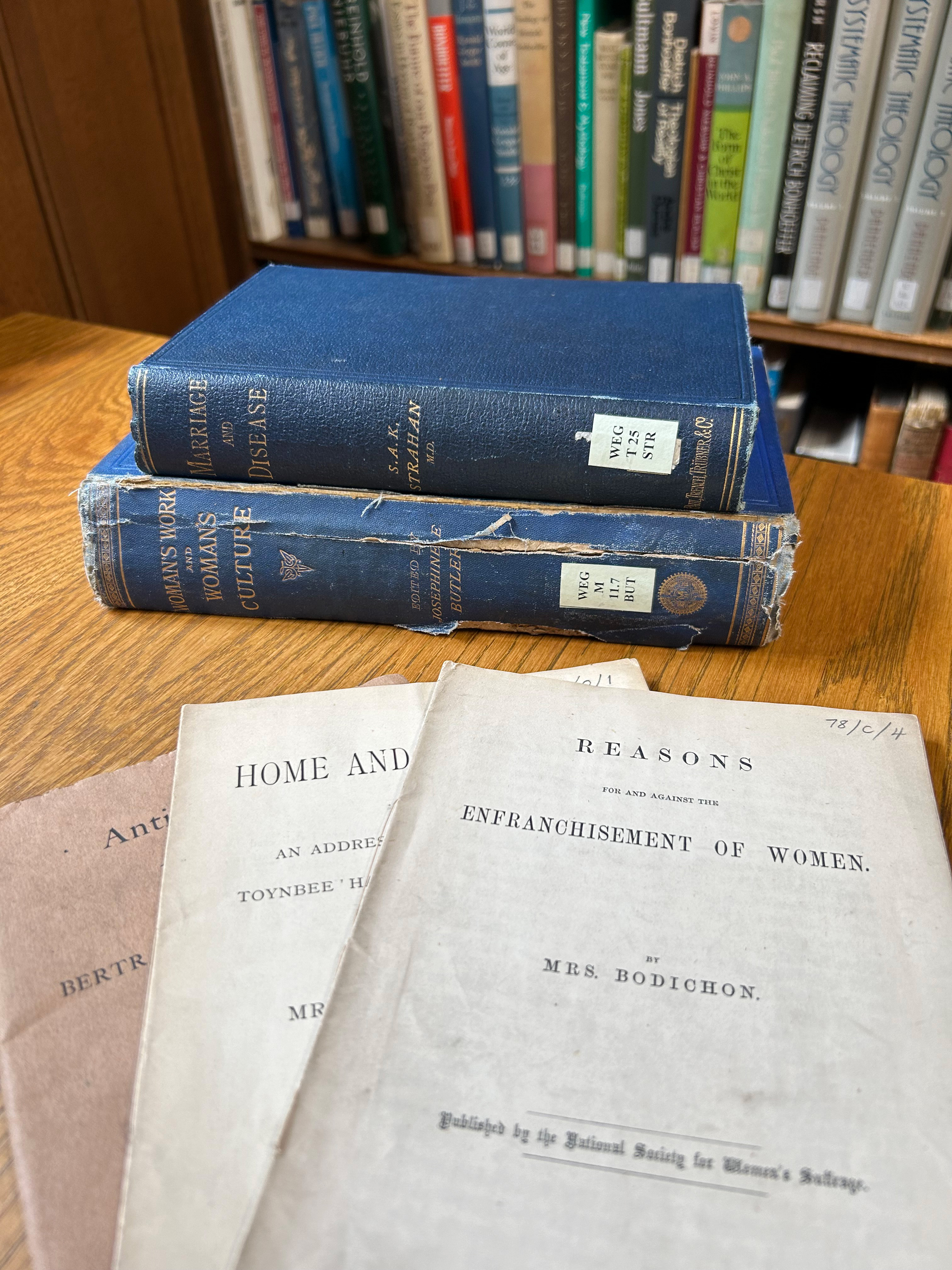 A pile of books on a desk, including 'Woman's work and woman's culture'. They are old books with blue cloth-bound spines. In the foreground there are pamphlets like 'The Enfranchisement of Women'