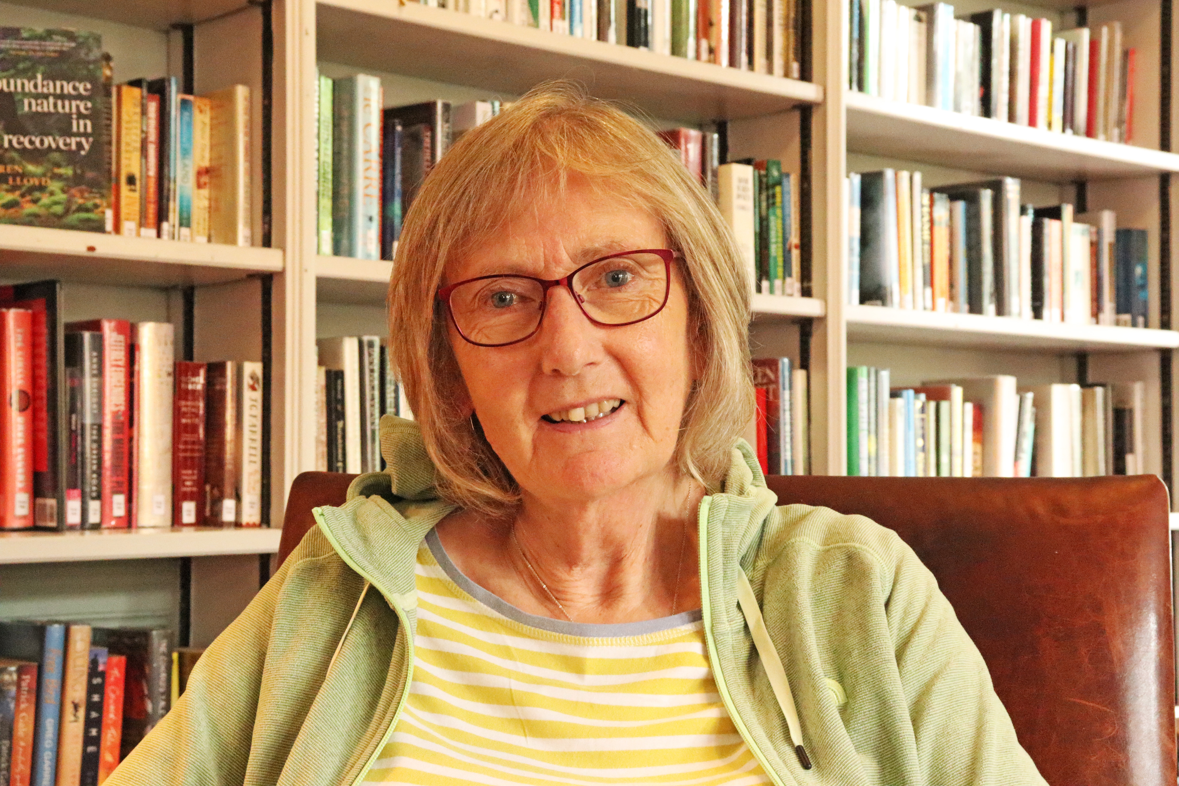 Author Karen Lloyd sitting in front of a bookcase in the Gladstone Room. Karen is wearing a hoodie style top and has blonde hair